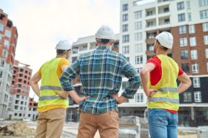 result back view three men protective helmets holding hands his waist standing construction site looking buildings construction during day