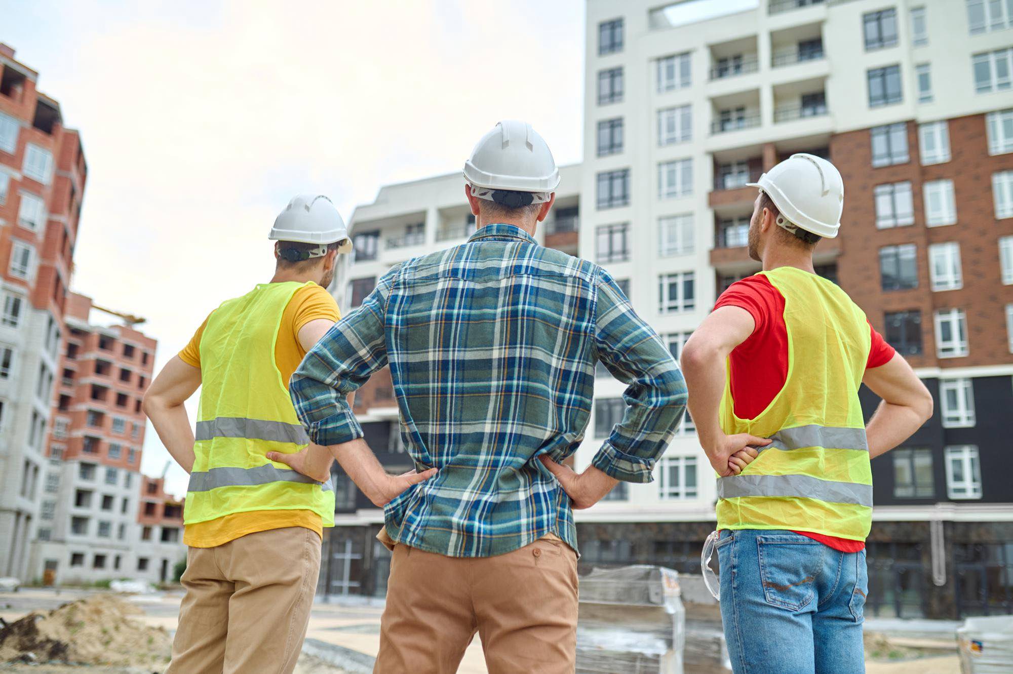result back view three men protective helmets holding hands his waist standing construction site looking buildings construction during day